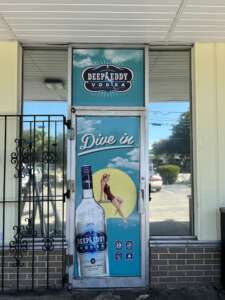 A door featuring an advertisement for Deep Eddy Vodka with an illustration of a woman next to a bottle and the text "Dive in," set against a background depicting the outdoors.