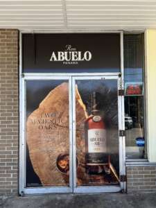 A storefront with doors displaying an advertisement for Ron Abuelo rum, featuring a bottle and wooden imagery. A "No Smoking" sign is also visible.