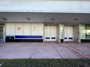 Storefront with closed metal shutters featuring a Walgreens logo and a pharmacy symbol. The pavement in front shows wear, and light reflects off the glass doors. Grass lines the edge of the sidewalk.