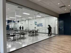 View through glass wall into a modern office space of Skeletal Dynamics with desks, chairs, and ceiling lights; four people are present inside and near the entrance.