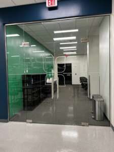 Glass-walled office with a green accent wall, chairs, and computer desks visible. Fluorescent ceiling lights are on, and there's an exit sign above the entrance. Trash can is near the doorway.