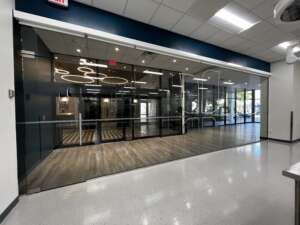 Glass entrance to a modern office or workspace with wood flooring, ceiling lights, and a sliding door. The interior shows a reception area and seating.