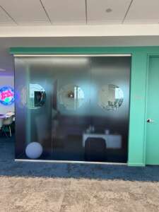 An office room with frosted glass doors featuring three circular designs, situated beside a teal-colored wall with a closed white door.