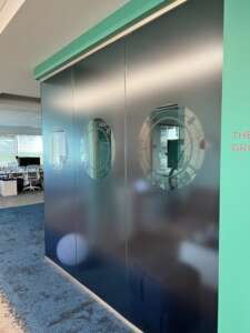 Office space with a dark-colored wall featuring circular windows. Visible in the background are desks, chairs, and computers through the windows. The wall has teal and white accents.