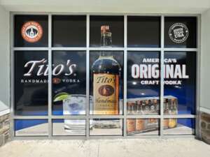 Storefront window advertisement for Tito's Handmade Vodka, featuring a large vodka bottle, a cocktail glass, and various promotional text, including "America's Original Craft Vodka.