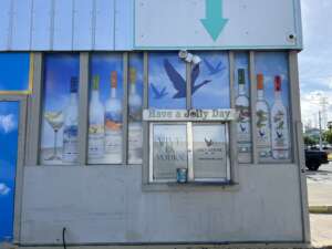 A storefront with Grey Goose vodka advertisements, including large bottle images and the slogan "Have a Jolly Day." A small window and door are also present. Blue sky and clouds in the background.