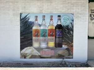 A wall advertisement displays four bottles of Cazadores tequila in different varieties in front of an agave field backdrop. A text bubble states "Made from 100% Blue Agave.