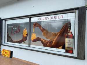 Storefront advertisement for Courvoisier featuring a bottle of VSOP cognac and two hands holding glasses with drinks. The text reads "Welcome to the Gala" and "Courvoisier.