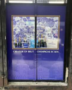 Storefront with blue signage displaying multiple bottles of Pommery champagne. Text reads "Creator of Brut Champagne in 1874." Background includes floral patterns and a historic building image.