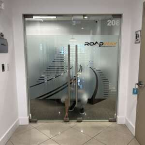 Glass doors with the name "RoadWay Construction, LLC" and office number 208. Inside, a person is reflected, crouching near a desk. Nearby is a bathroom door with a men's restroom sign.