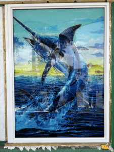 A framed painting of a leaping marlin, vibrant in blues and whites, displayed outdoors. The marlin is depicted breaking through the water's surface against a backdrop of a setting sun.