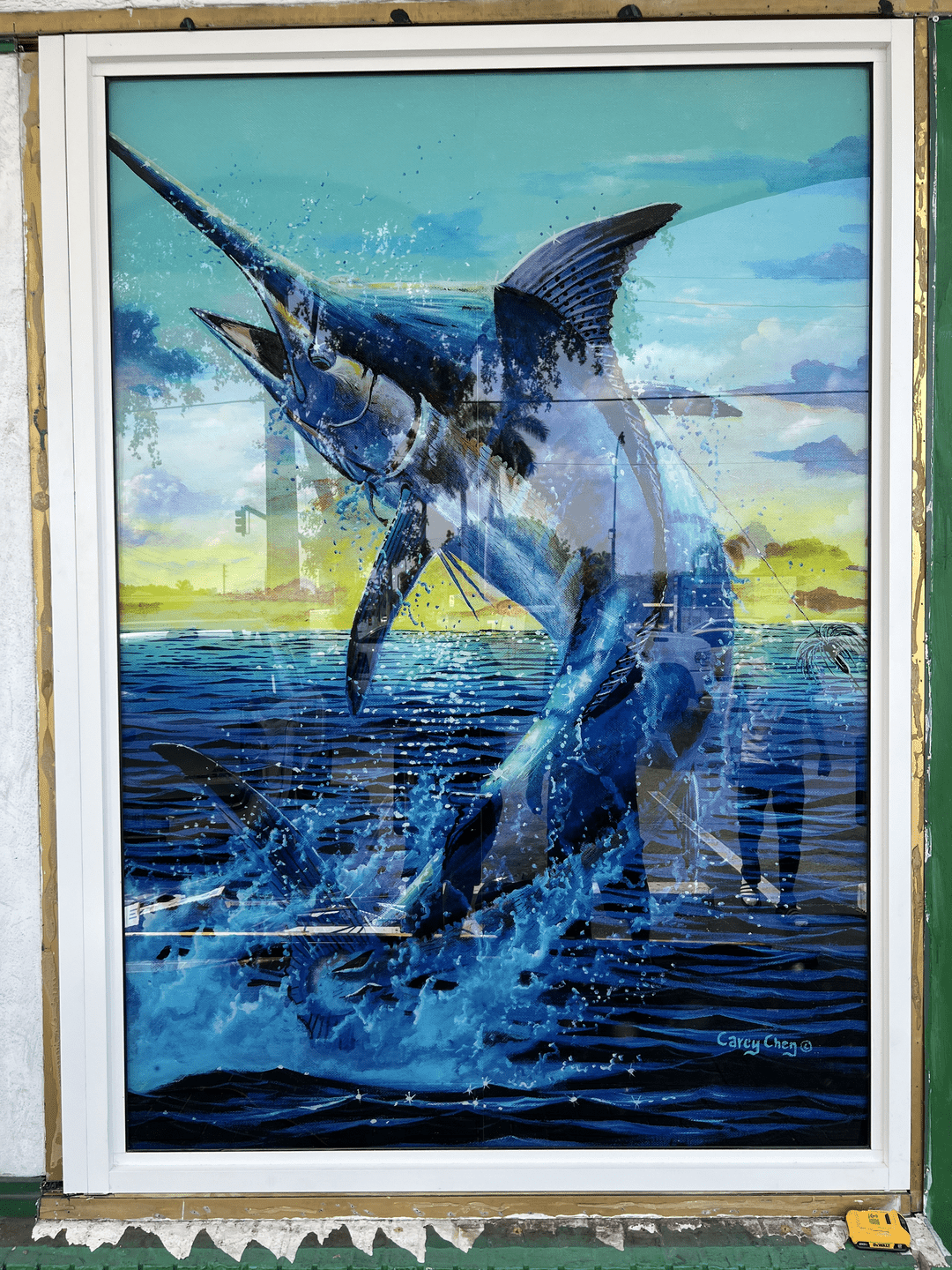 A framed painting of a leaping marlin, vibrant in blues and whites, displayed outdoors. The marlin is depicted breaking through the water's surface against a backdrop of a setting sun.